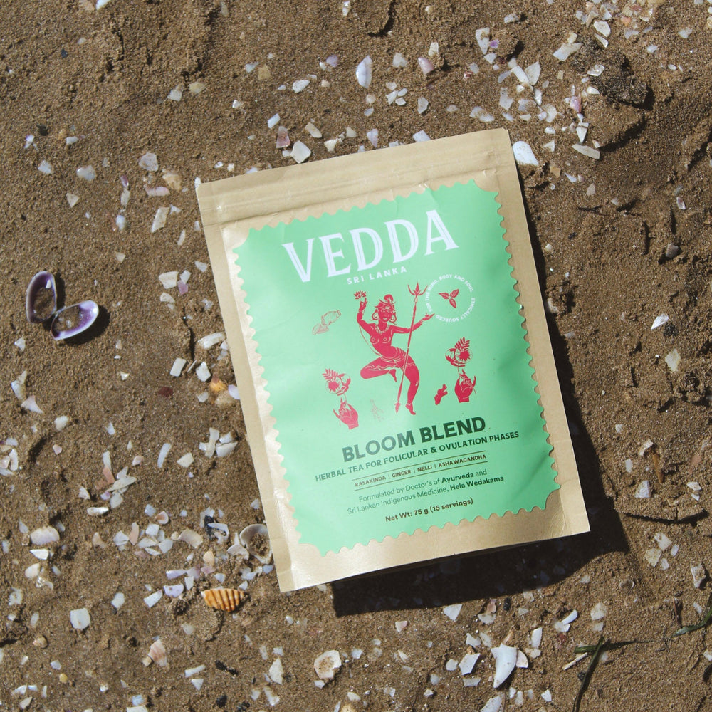 A kraft paper pouch of VEDDA Tea 'Bloom Blend' herbal tea lies on sandy ground scattered with small white shells. The pouch has a light green label with a red dancer illustration and black text.