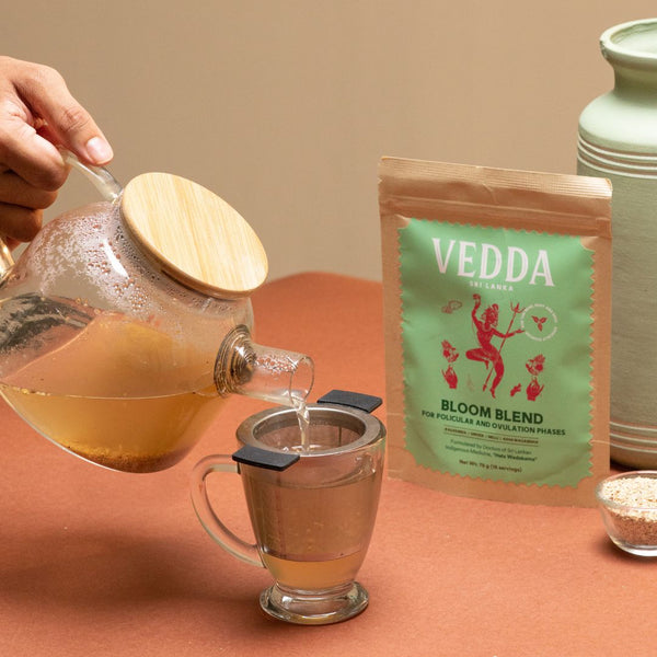 A hand pours tea from a glass teapot with a wooden lid into a glass mug with a metal filter. A stand-up pouch of VEDDA Botanicals Bloom Blend tea stands next to it on a terracotta surface against a warm beige background.