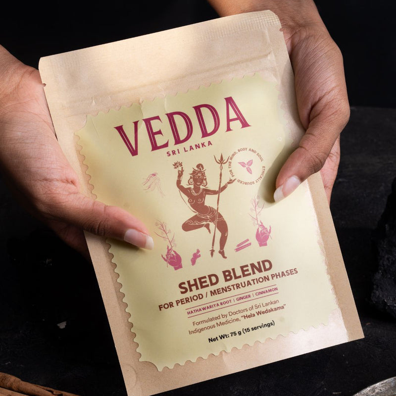 A person holds a stand-up pouch of VEDDA Sri Lanka 'Shed Blend' herbal medicine. The pouch is a warm cream color with dark red text and an illustration of a dancing figure.