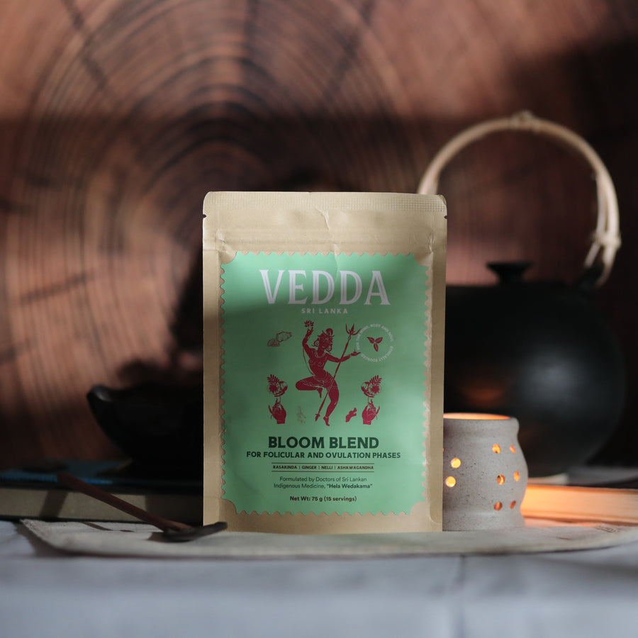 A stand-up pouch of VEDDA Satva Bloom Blend herbal tea placed on a table, with a black teapot and a small lit candle in the background against a dark wood grain backdrop.