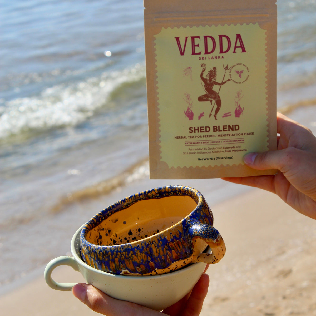 A person at the beach holds a cup of tea in one hand and a package of VEDDA Shed Blend tea in the other, with the ocean and sandy shore in the background.