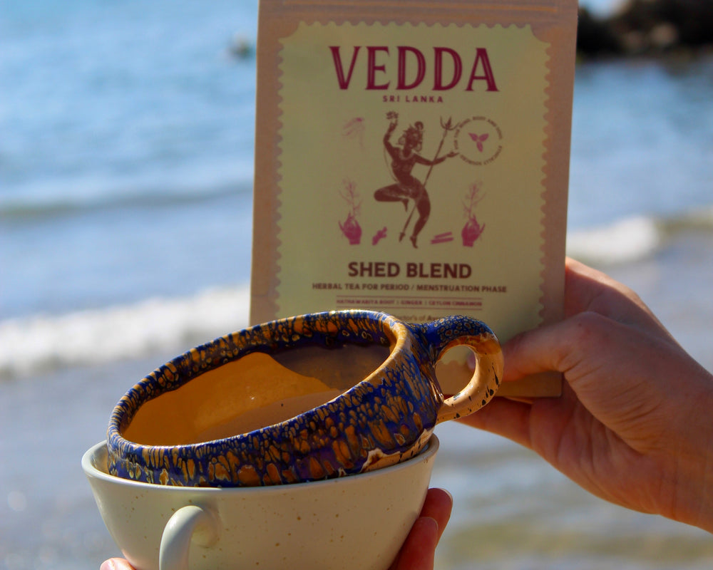 A person holds a white ceramic mug with a blue patterned inner cup at the beach, with a pouch of VEDDA Shed Blend coffee standing behind them.