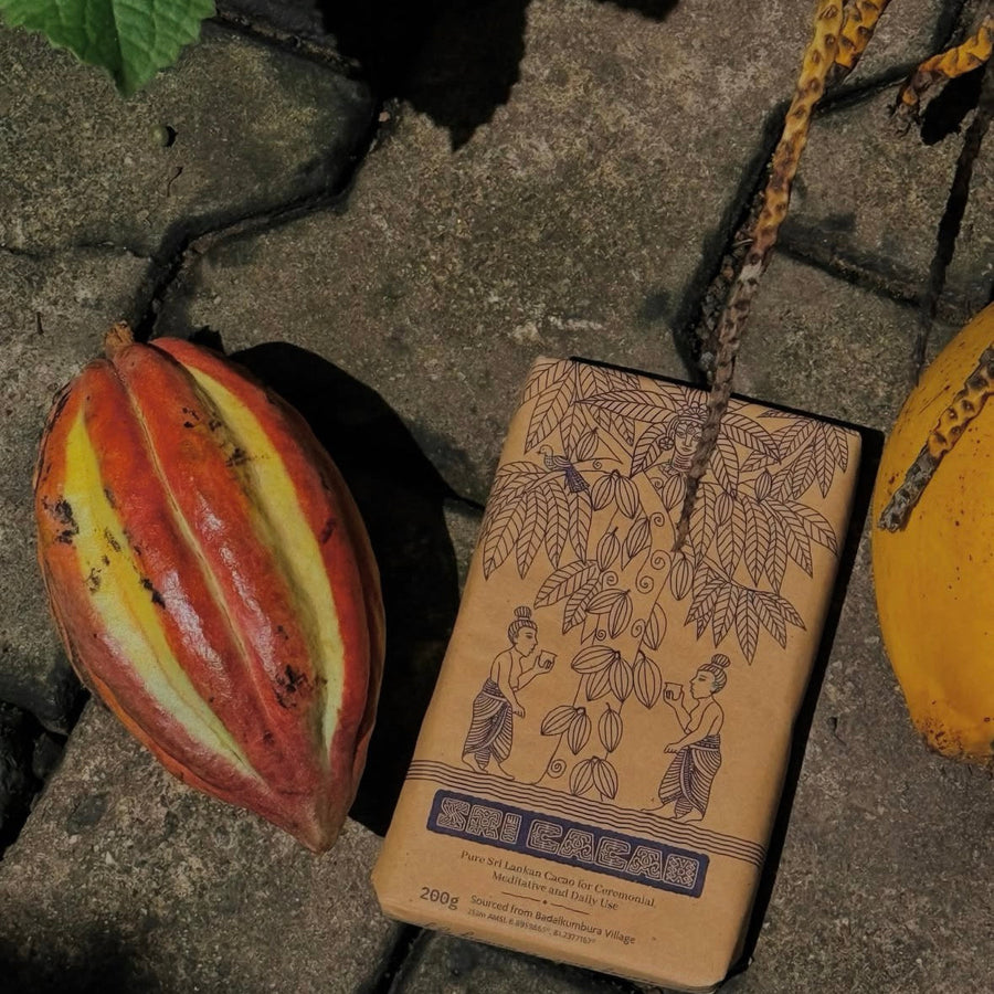 A package of Sri Lanka organic cocoa beans lies on a stone surface next to a ripe red and yellow cacao pod and a yellow mango, with a leaf in the top left corner.